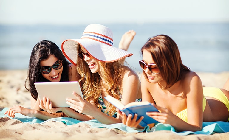 tre ragazze sulla spiaggia che leggono e guardano un tablet